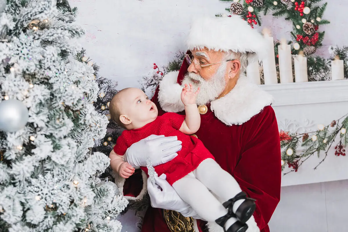 Santa holding baby by Christmas tree.